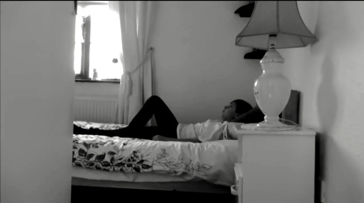 Black-and-white view through a doorway of a performer reclining on a bed, with a bedside lamp in the foreground.