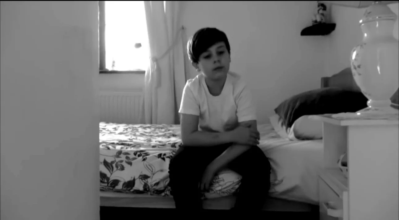 A young performer sitting on the edge of a bed, framed by the bedroom doorway.